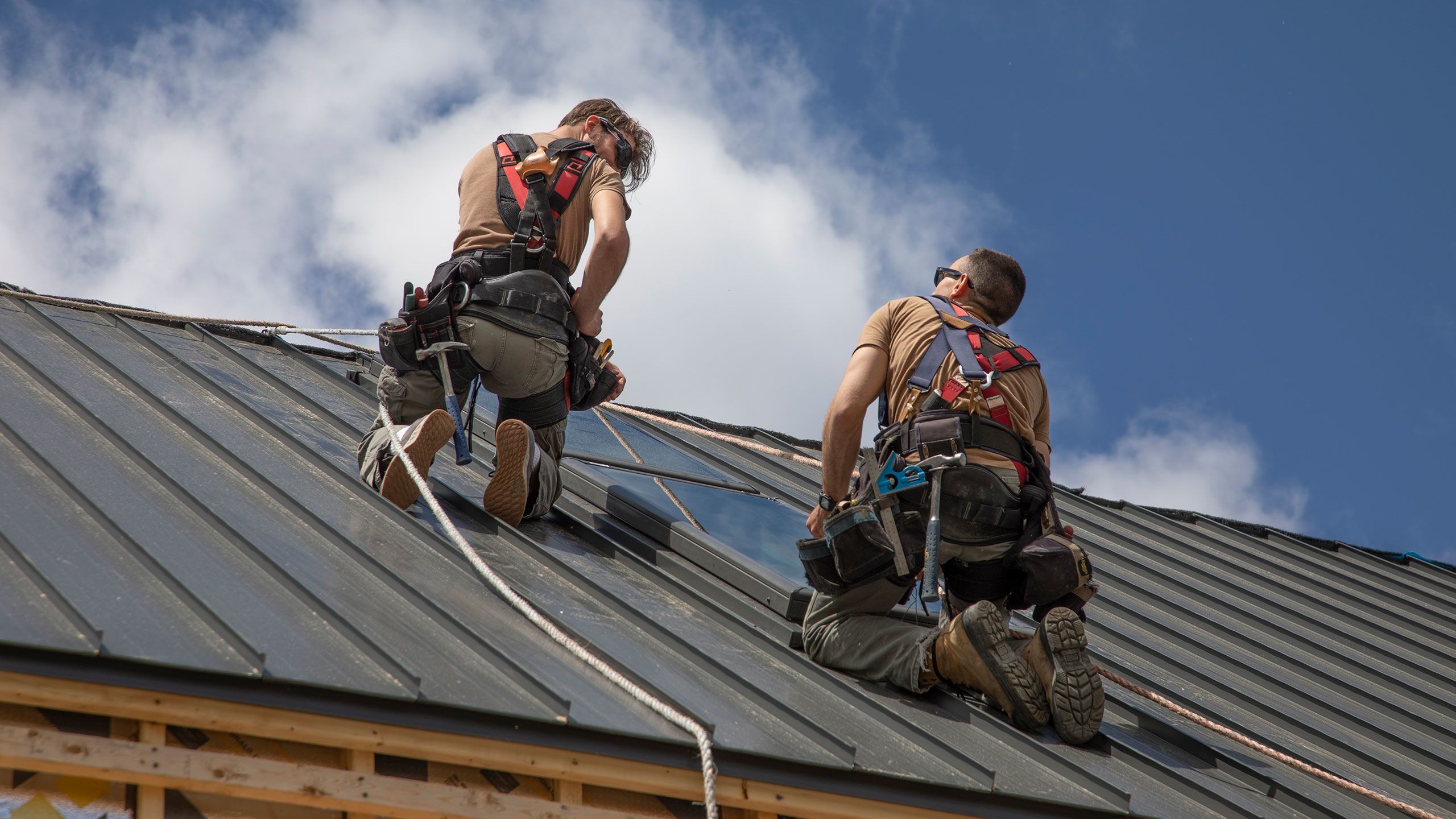 Two roofers tied off with ropes work installing the steel panels on the steeply pitched roof