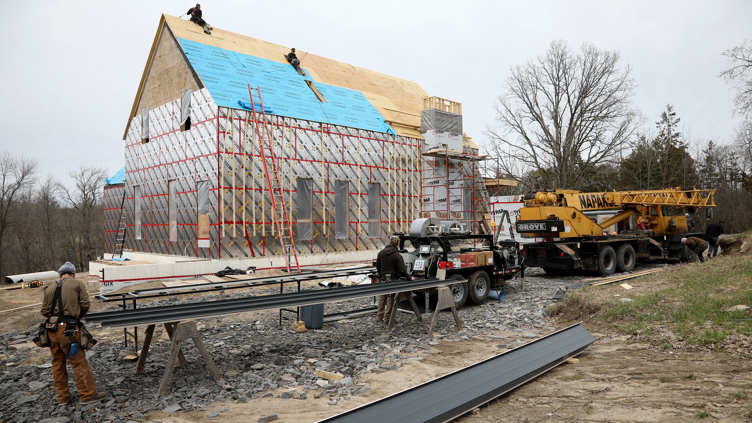 Roofers use a bending machine to create the standing seam steel panels for the roof. In the background other roofers install an installation layer