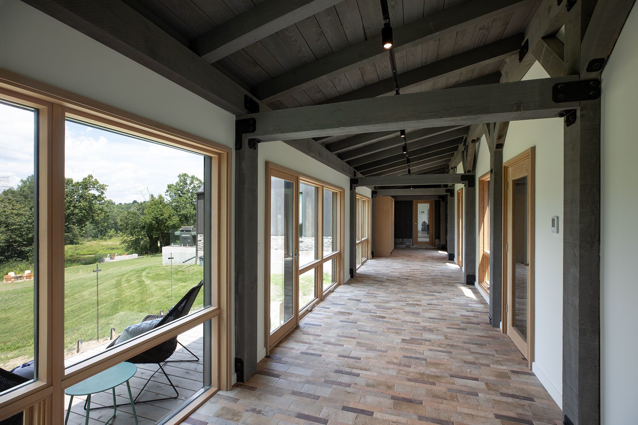 Walkway with gray stained timber frame roof, large windows and brick coloured tile floor. 